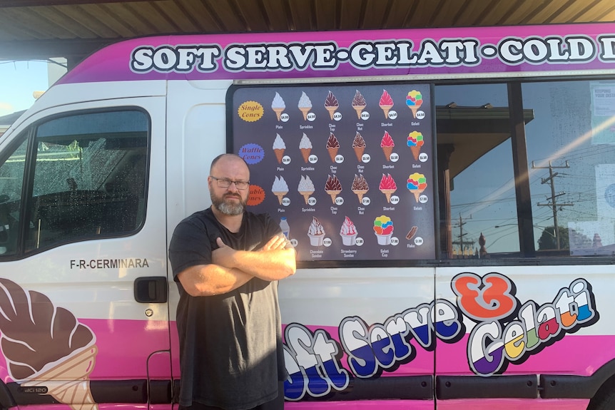 A middle aged man with a beard, glasses and black t-shirt stands in front of an ice cream van with his arms crossed