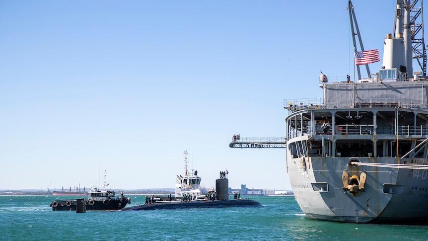 The top bit of a submarine in the water next to a large grey ship flying the US flag