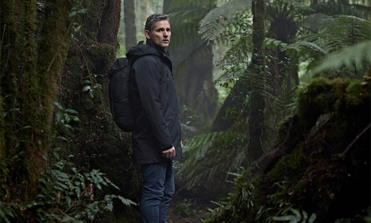 Eric Bana, in character as Aaron Falk, stands in jeans and a warm coat, wearing a backpack on a hiking trail in the bush.