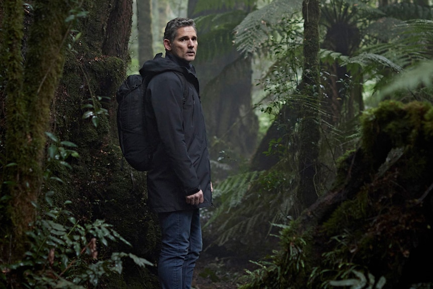 Eric Bana, in character as Aaron Falk, stands in jeans and a warm coat, wearing a backpack on a hiking trail in the bush.
