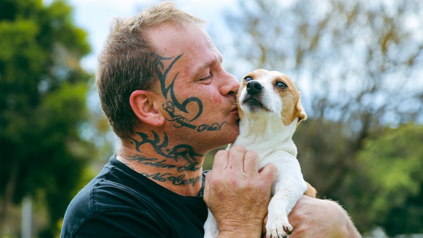 Brian Warton, a man with face and neck tattoo, holds a small dog and gives it a kiss on its cheek.