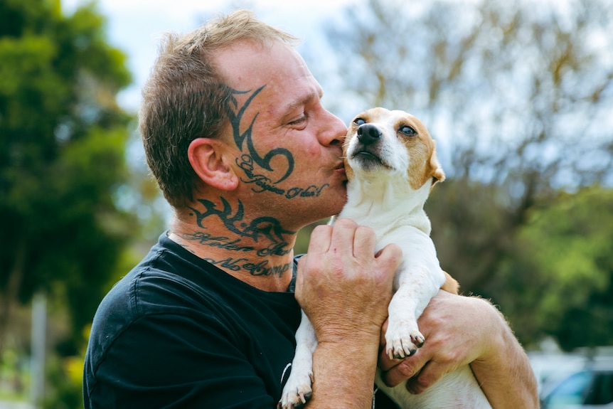 Brian Warton, a man with face and neck tattoo, holds a small dog and gives it a kiss on its cheek.