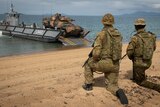 Armoured Army vehicle drives off boat onto the beach while two soldiers watch on 