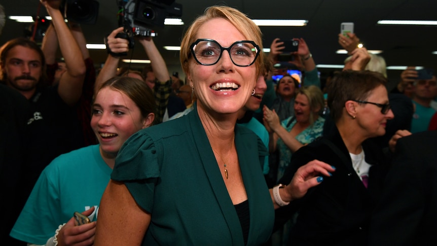 A woman wearing a teal shirt and black glasses smiles amid a sea of people. 