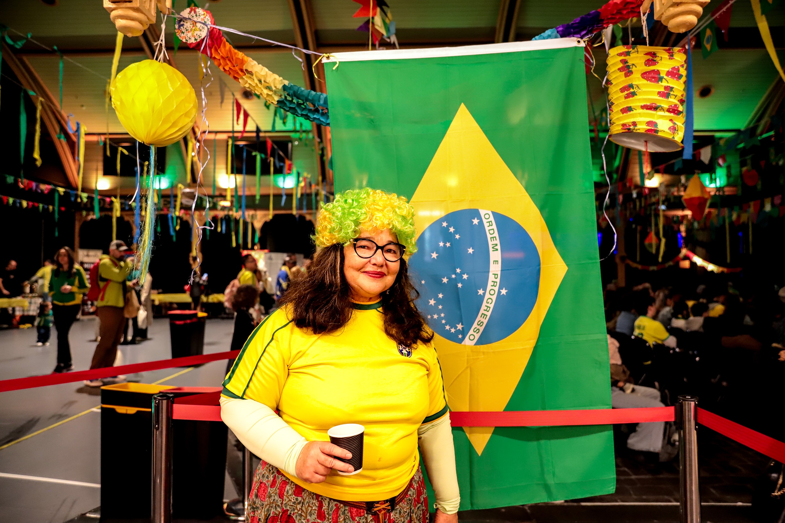 Woman wearing yellow Brazil shirt with yellow and green wig stands in front of a Brazillian flag with a lively event behind her