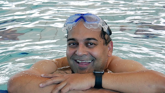 A middle-aged man wearing goggles smiles at the edge of a swimming pool