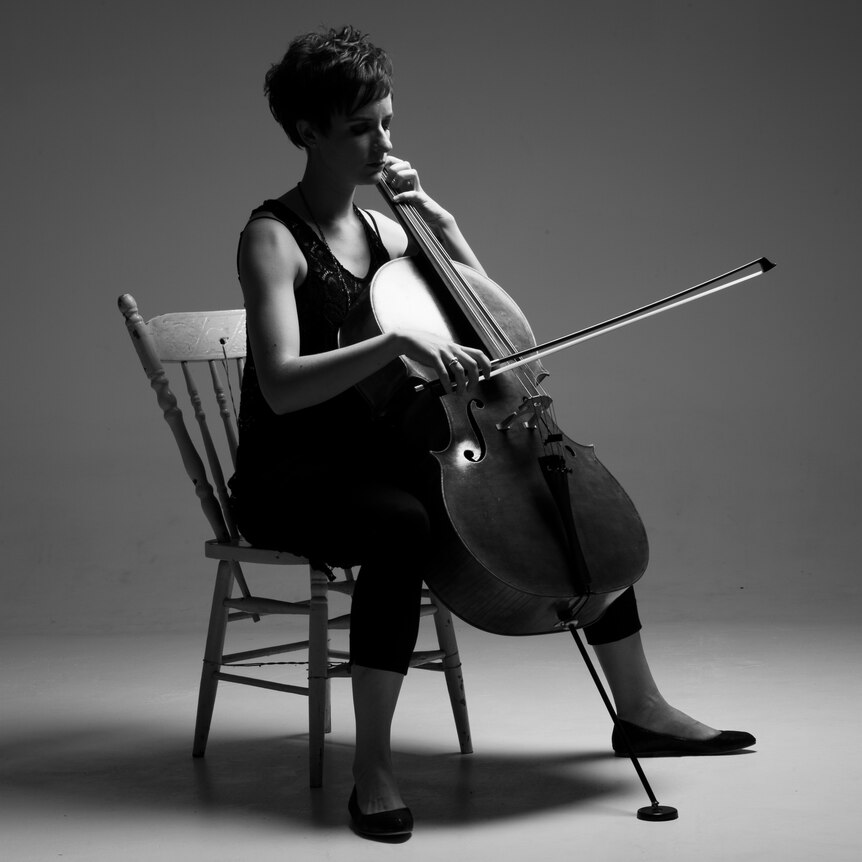Jamie Mills is sitting on a wooden chair playing her cello, looking downwards.