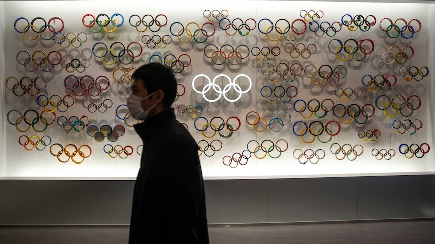 A man wearing a face mask walks past a lit display with dozens of versions of the Olympic rings.