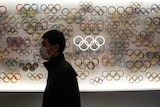 A man wearing a face mask walks past a lit display with dozens of versions of the Olympic rings.