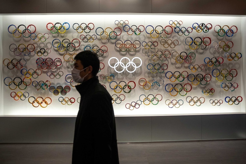 Un hombre que lleva una máscara facial pasa junto a una pantalla iluminada con docenas de versiones de los anillos olímpicos.