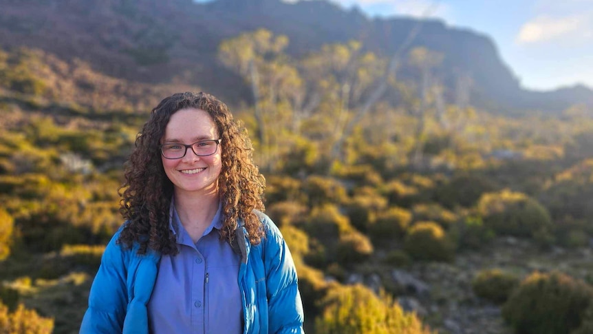 a woman in a blue jacket is looking at the camera smiling, she is in the bush and there is a mountain in the background
