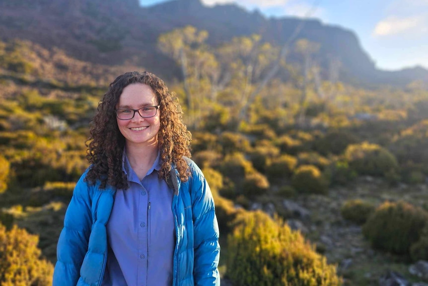 a woman in a blue jacket is looking at the camera smiling, she is in the bush and there is a mountain in the background