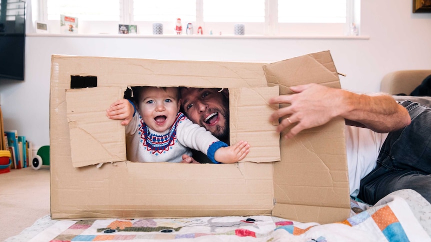 A small toddler pokes his head out of a cardboard box, happily playing with his father.