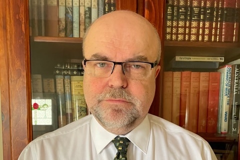A man in a formal shirt and tie stands in front of a bookcase.