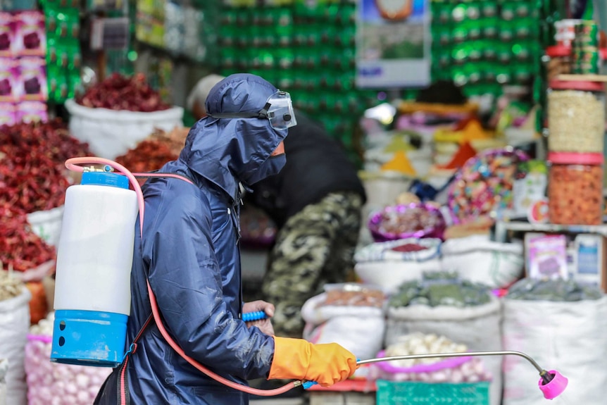 A man in protective gear spraying liquid near an Indian spice market