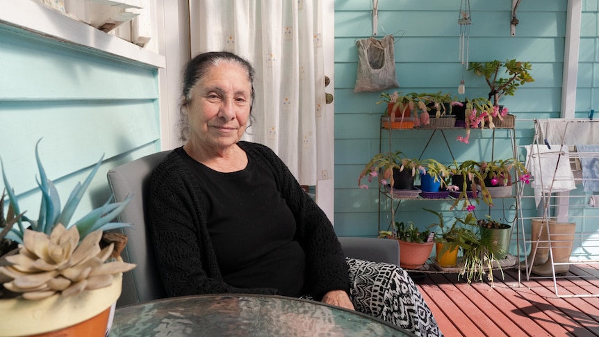 An elderly woman sits in the sun on her back deck, smiling.