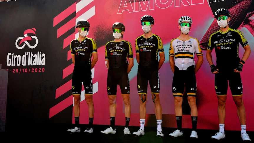 A group of masked cyclists stand next to each other on a stage next to a Giro d'Italia sign.