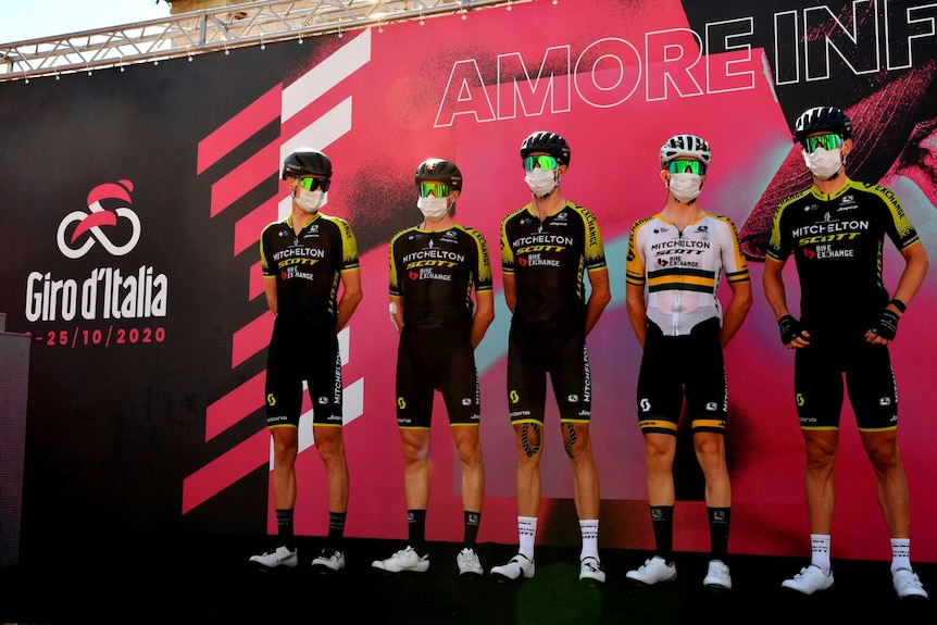 A group of masked cyclists stand next to each other on a stage next to a Giro d'Italia sign.