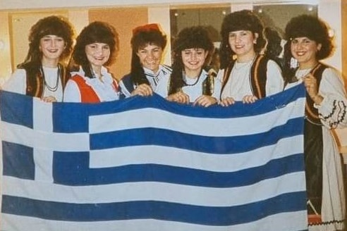 Six women wearing Greek traditional costume, holding a Greek flag in front of them. 