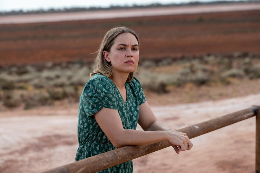 A young Indigenous woman with light brown hair wears a green dress and leans on a rusty fence in a dusty landscape.