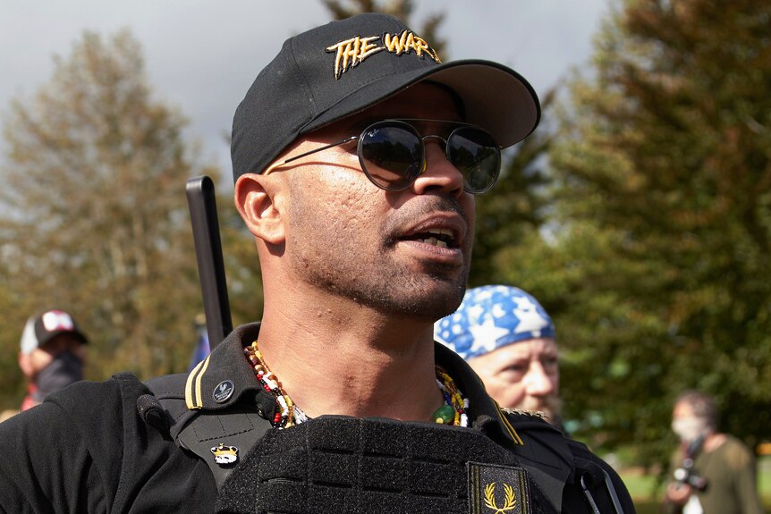 A close up of Enrique at a rally outside wearing black sunglasses and a black cap that says The War.