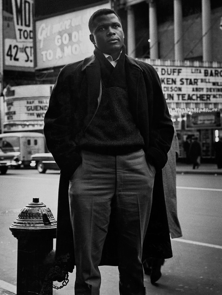 Black and white image of Sidney Poitier standing in a NYC street in front of billboards