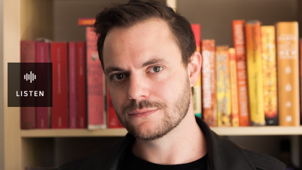 Young man looking at camera with book shelf behind him. Has Audio.