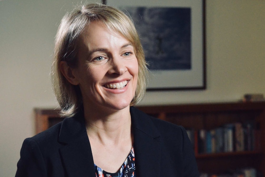 Dr Kemp smiles in her home. A picture and book case is out of focus behind her.