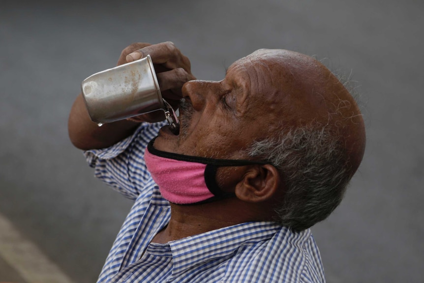 A bald man with a pink face mask lowered to his chin drinks clear water from a small discoloured metal cup.