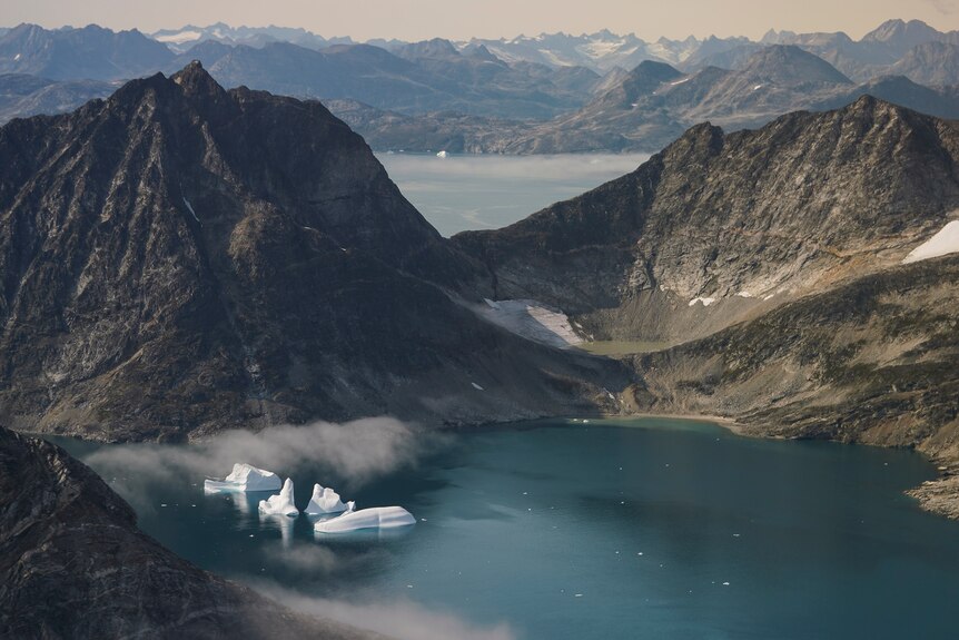 Four icebergs sit near each other on a lake of water surrounded by mountains. Birds eye view. 