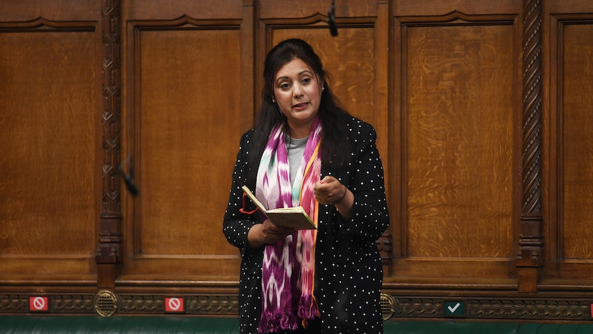 A middle-aged Indian woman in black jacket and pink scarf holds a book and speaks in ornate room