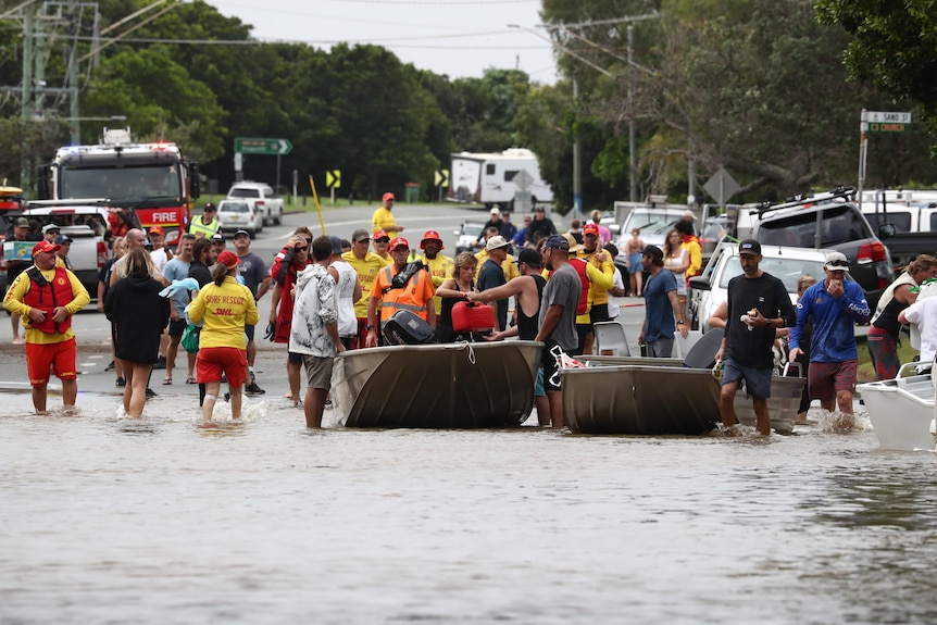 A crowd of people wade through floodwaters and sit on boats