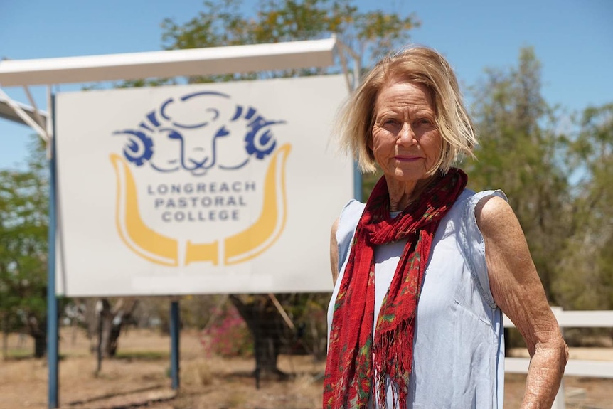 Woman with blonde hair, wearing a white top, red scarf and standing in front of a pastoral college sign