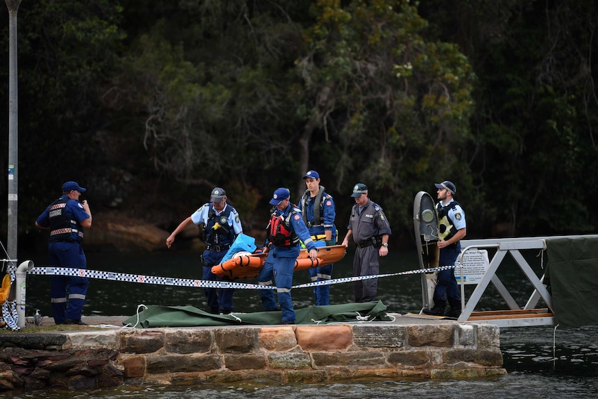 The body of a seaplane passenger is covered in a blue sheet and carried on a stretcher by four police officers.