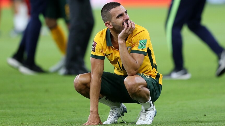 A male soccer player wearing yellow and green crouches down on the grass after a game