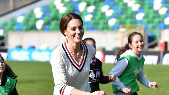 Kate wears a white, red and blue jumper and black leggings as she runs on field with teenage girls behind her. She is smiling.