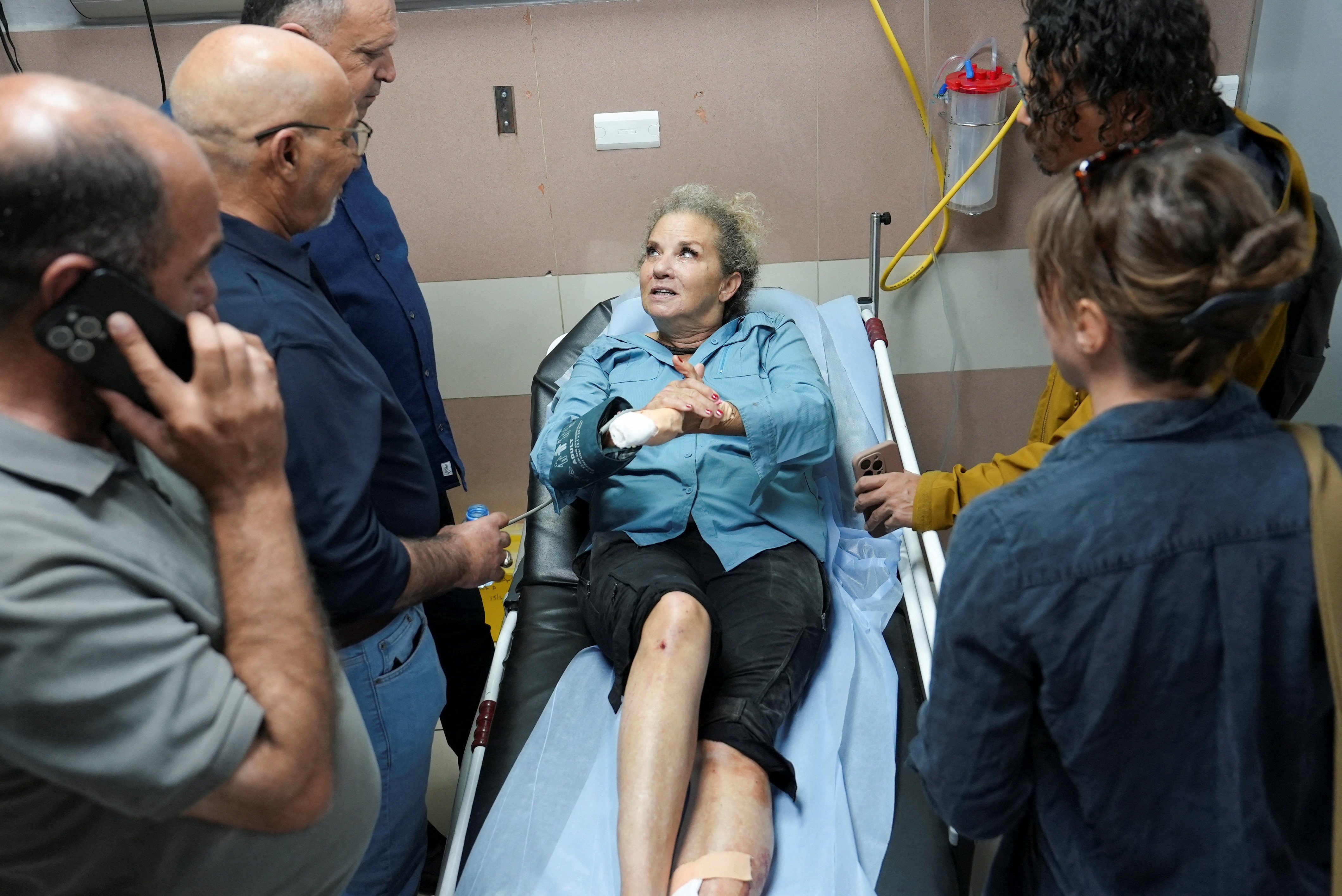 A blonde woman with a hand in bandages talks to a group of people surrounding her hospital bed.