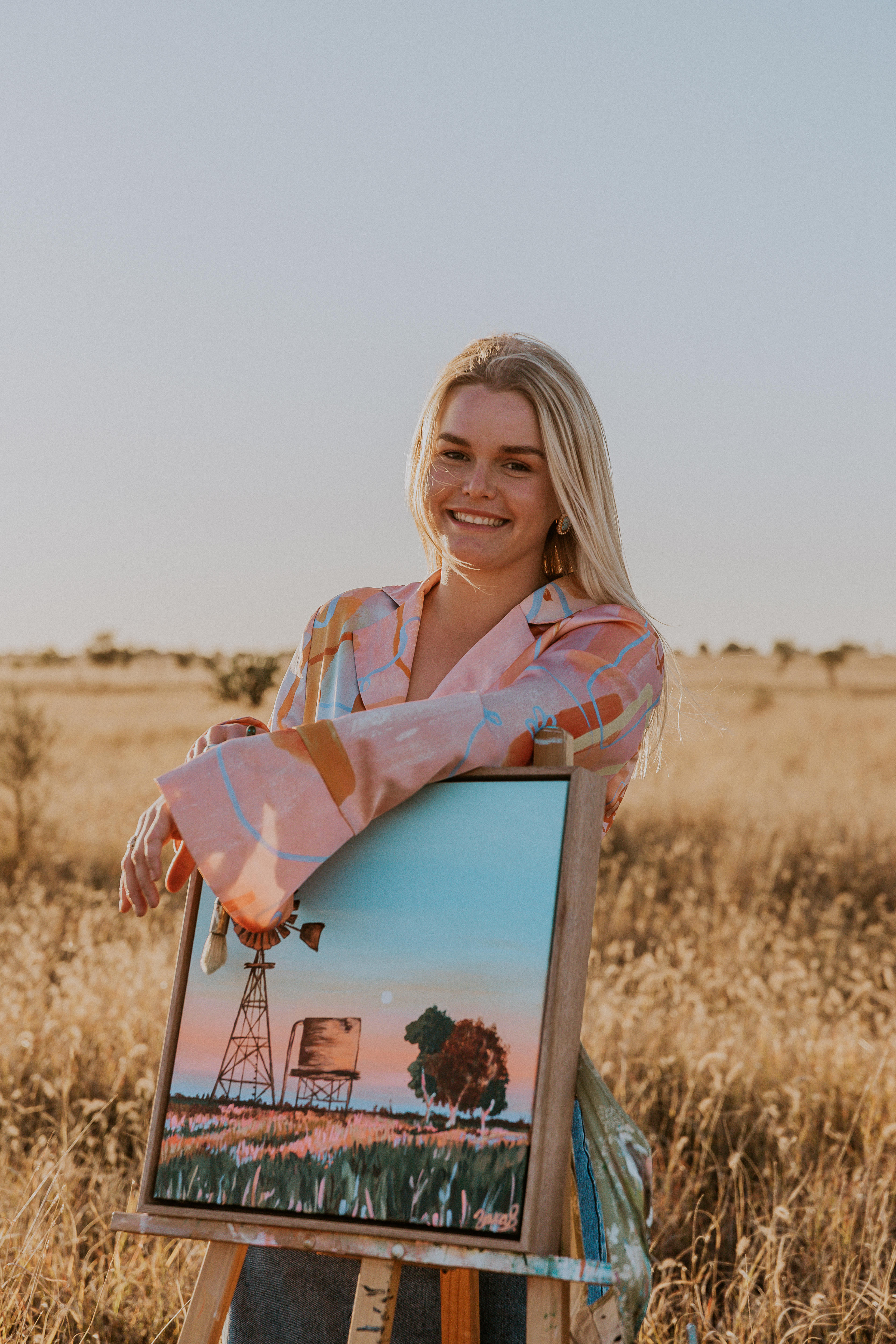 Zarabella posa para uma foto profissional apoiada em uma de suas pinturas em um campo.  