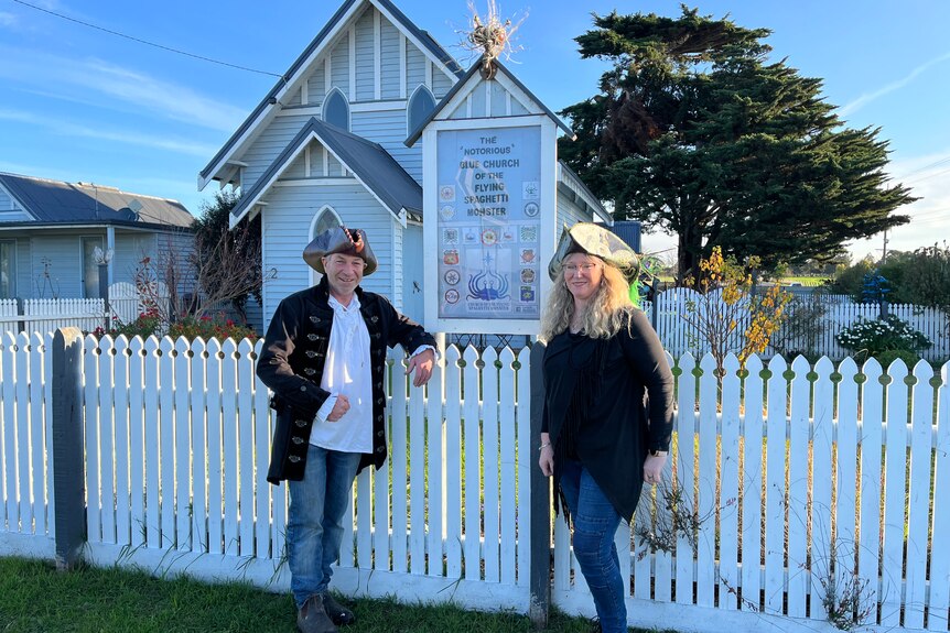 Two people wearing pirate hats stand outside a small country church.