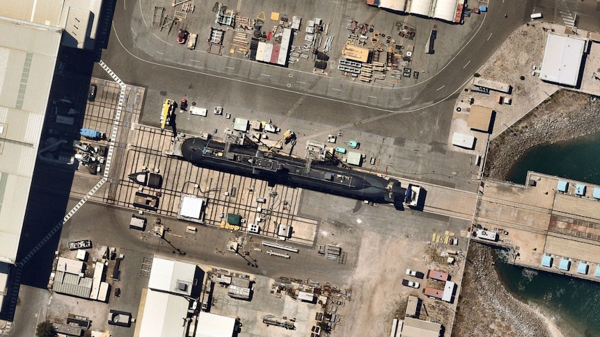A satellite image of a large black submarine on land surrounded by industrial buildings and cranes near the water.