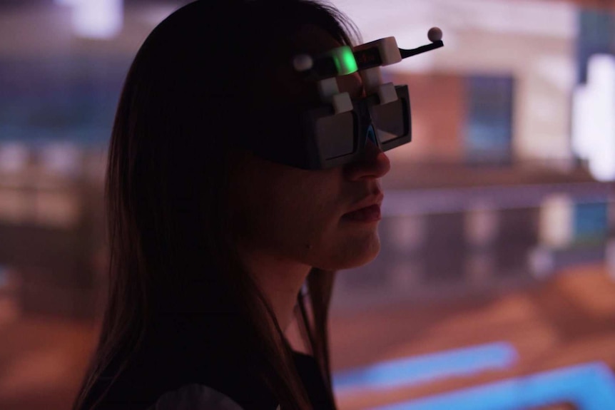 A young woman with brown hair wears a virtual reality headset as she stands in a dark room with bright screens behind her.