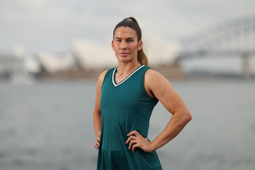Australian rugby sevens player Charlotte Caslick stands on a rock in front of SydneyHarbour in a green Olympic Games uniform.