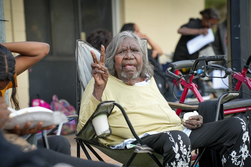 Elderly Indigenous woman with grey hair sit on a camping chair and makes a peace sign with her fingers