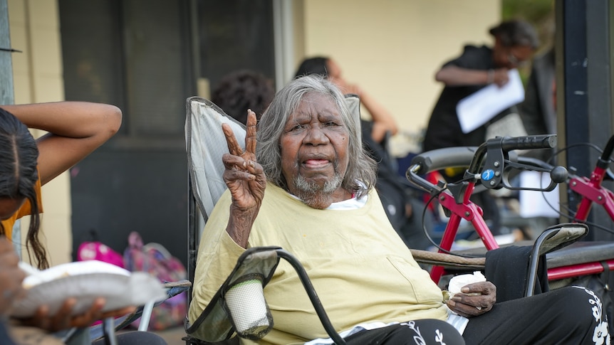 Elderly Indigenous woman with grey hair sit on a camping chair and makes a peace sign with her fingers