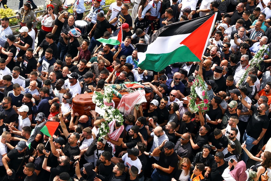 Thousands walk along funeral procession, with Palestinian flag flying overhead.