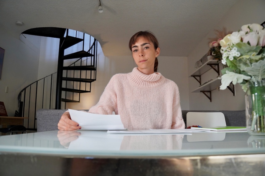 Sarah Kay wearing a pale pink turtleneck jumper, holding a document at her kitchen table.