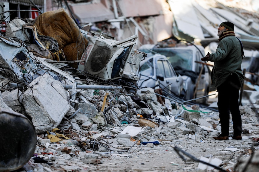 A man stands next to a destroyed building that has been reduced to rubble. 