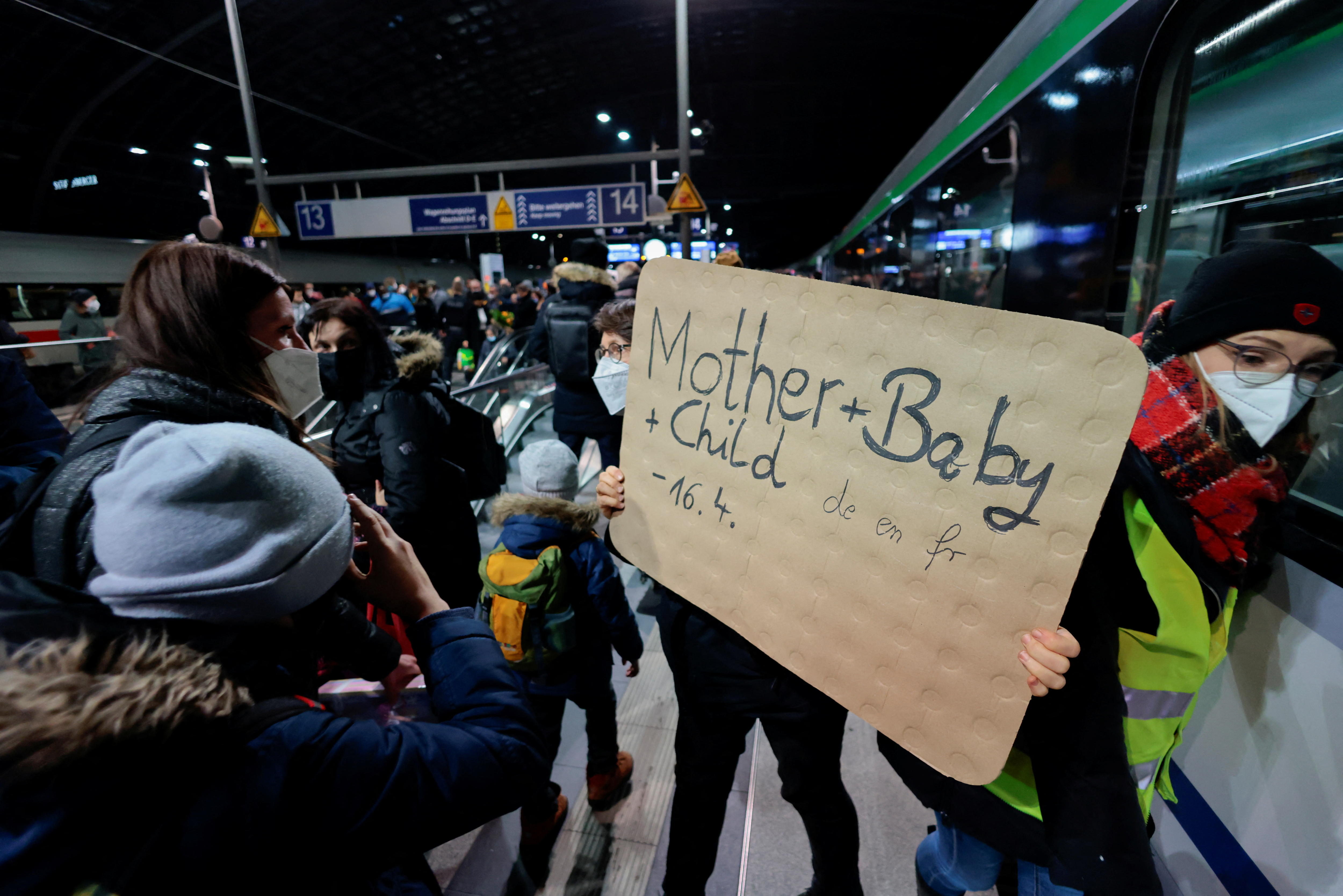Person at busy train station holds sign offering accommodation for mother, baby and child.