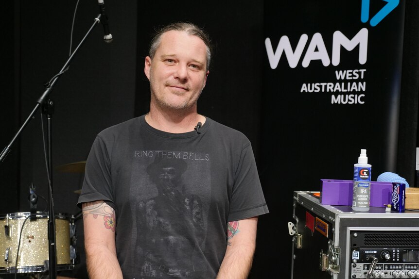 A man in a black shirt sitting next to a sign which says WAM West Australian Music 