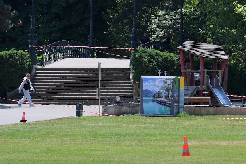A woman in white forensic clothing walks in a cordoned-off park next to a playground.